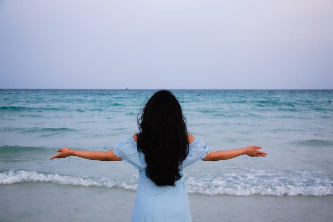 A woman stands at the ocean's edge with arms outstretched, embodying restored balance and renewed sense of freedom — reflecting the transformative outcomes of upper cervical care for vertigo.