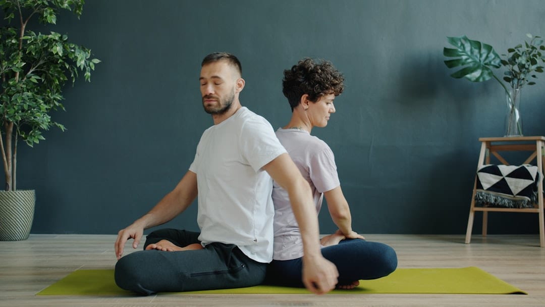 Two people sitting back-to-back on a yoga mat in a calm wellness center, practicing mindful posture exercises that support spinal health and upper cervical care.