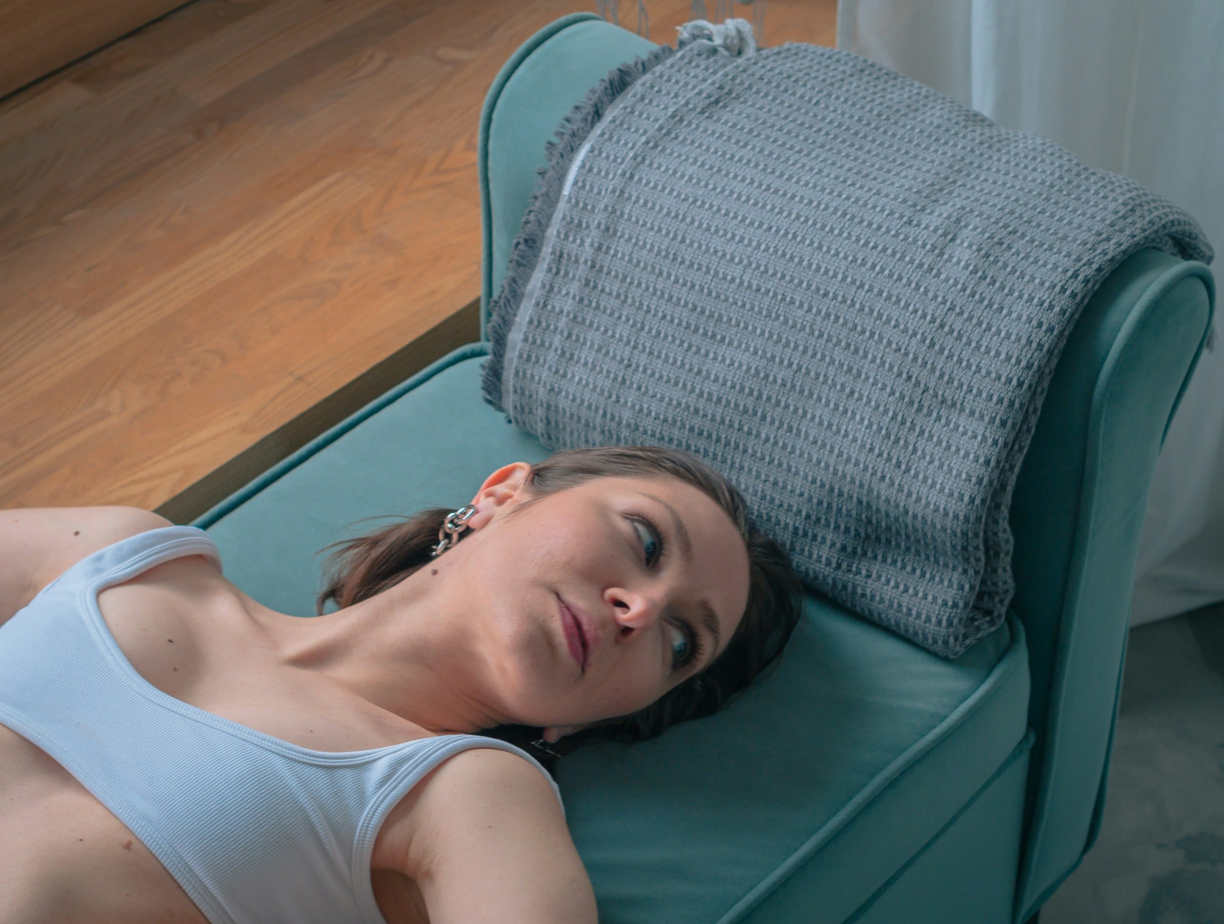 A woman resting with her head back on a teal couch with a soft gray pillow, taking a quiet moment to recover from a headache at home.
