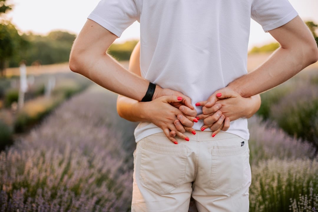 A person standing in a sunlit field with hands resting on their lower back, representing the everyday impact of lower back discomfort and the path toward restored function with upper cervical care.