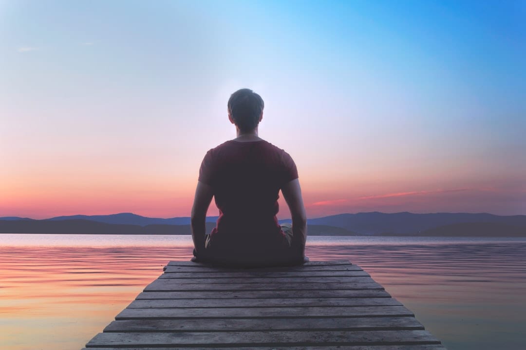 A person seated in a calm meditative pose on a dock at sunset, reflecting the mental clarity and nervous system restoration that upper cervical care supports.