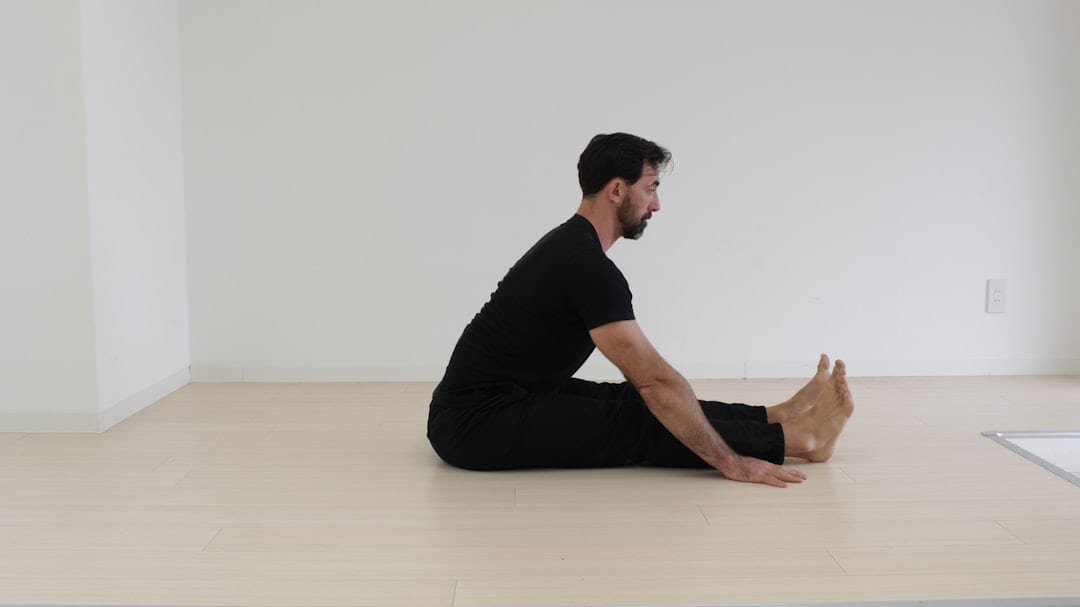 A man seated on the floor with legs extended, gently stretching — a calm, relatable image representing the discomfort and recovery journey associated with sciatica and upper cervical care.