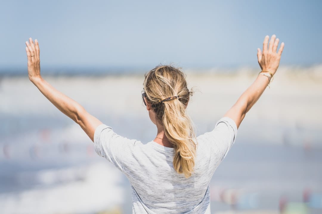 A woman stands at the beach with arms raised openly, embodying the restored mobility and renewed vitality that upper cervical care can provide for those recovering from sciatica.