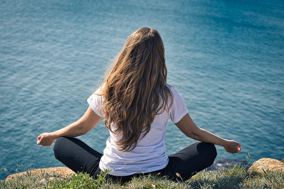 A woman seated in a calm, meditative pose overlooking a serene blue sea, reflecting the restored function, mental clarity, and whole-body wellness that upper cervical care supports.